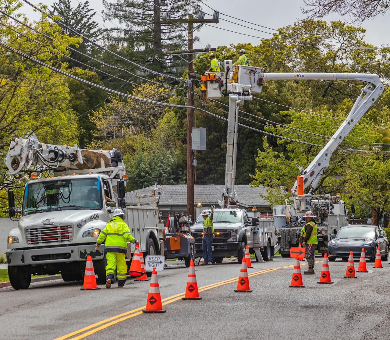 about-07 Utility workers in reflective gear repair power lines using bucket trucks and cones on a suburban street.